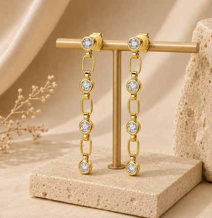 Liora gold linear drop earrings with linked sparkling round stones displayed on a beige stone stand.