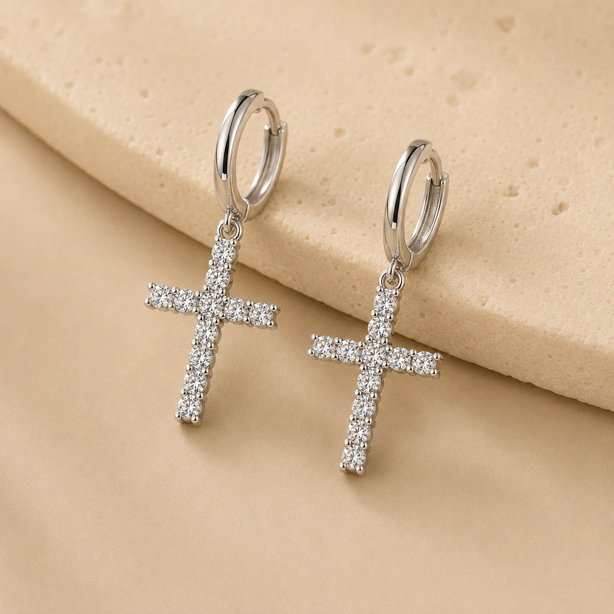 Dainty silver-tone cross drop hoop earrings with sparkling moissanite stones on a beige surface.