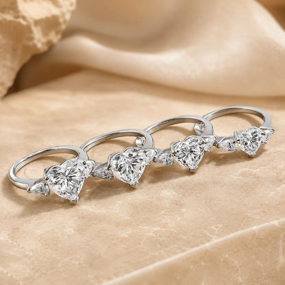 Heart-cut three-stone moissanite engagement rings displayed in multiple center-stone sizes on a warm beige surface.