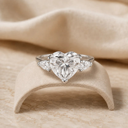 Heart-cut moissanite engagement ring with two sparkling side stones displayed on a soft beige jewelry stand.