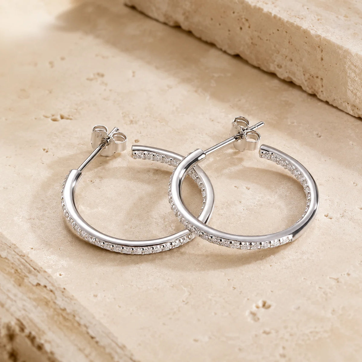 Isadora™ 925 sterling silver moissanite hoop earrings resting on a beige stone surface with secure post backs.