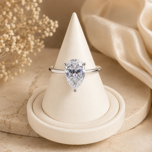 Pear-cut moissanite engagement ring displayed upright on a minimalist beige jewelry stand with soft luxury styling.