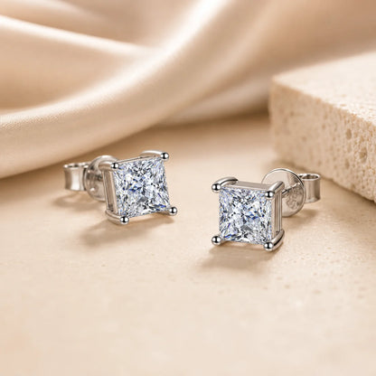 Close-up of princess-cut moissanite stud earrings with sparkling square stones in polished silver four-prong settings.