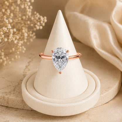 Rose gold pear-cut moissanite engagement ring featuring a brilliant solitaire center stone displayed on a soft beige jewelry stand.