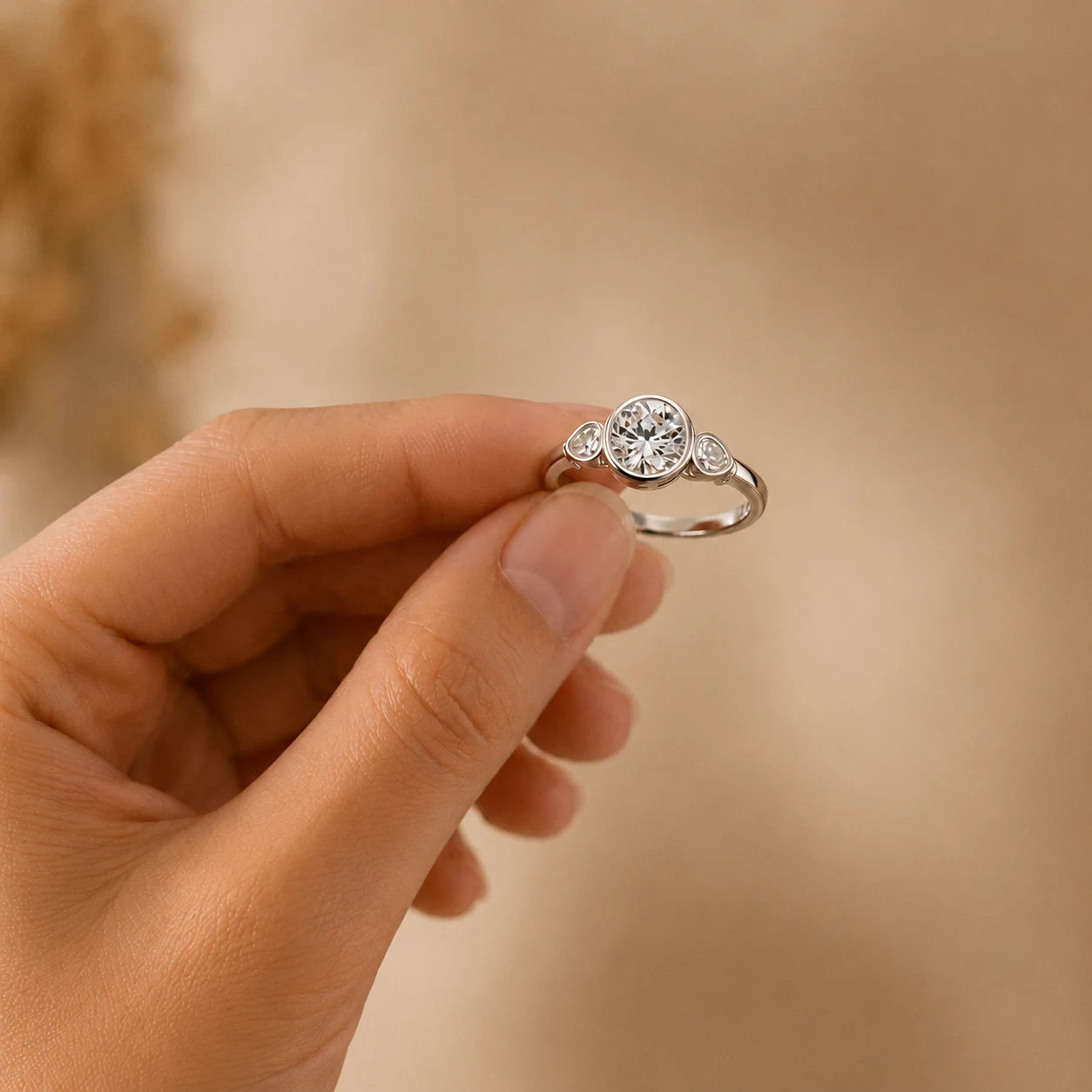 Close-up of a silver round moissanite three-stone ring held by hand against a warm beige background.