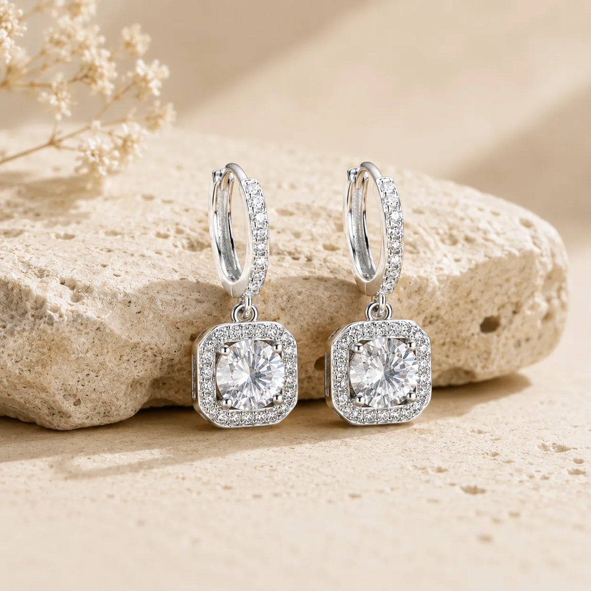 Marielle™ silver halo moissanite drop earrings displayed on a textured beige stone surface.
