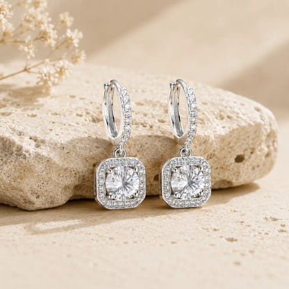 Marielle™ silver halo moissanite drop earrings displayed on a textured beige stone surface.