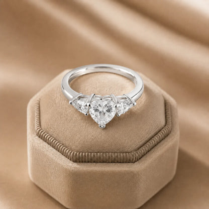 Silver heart-cut three-stone moissanite engagement ring displayed on an elegant beige velvet jewelry box.