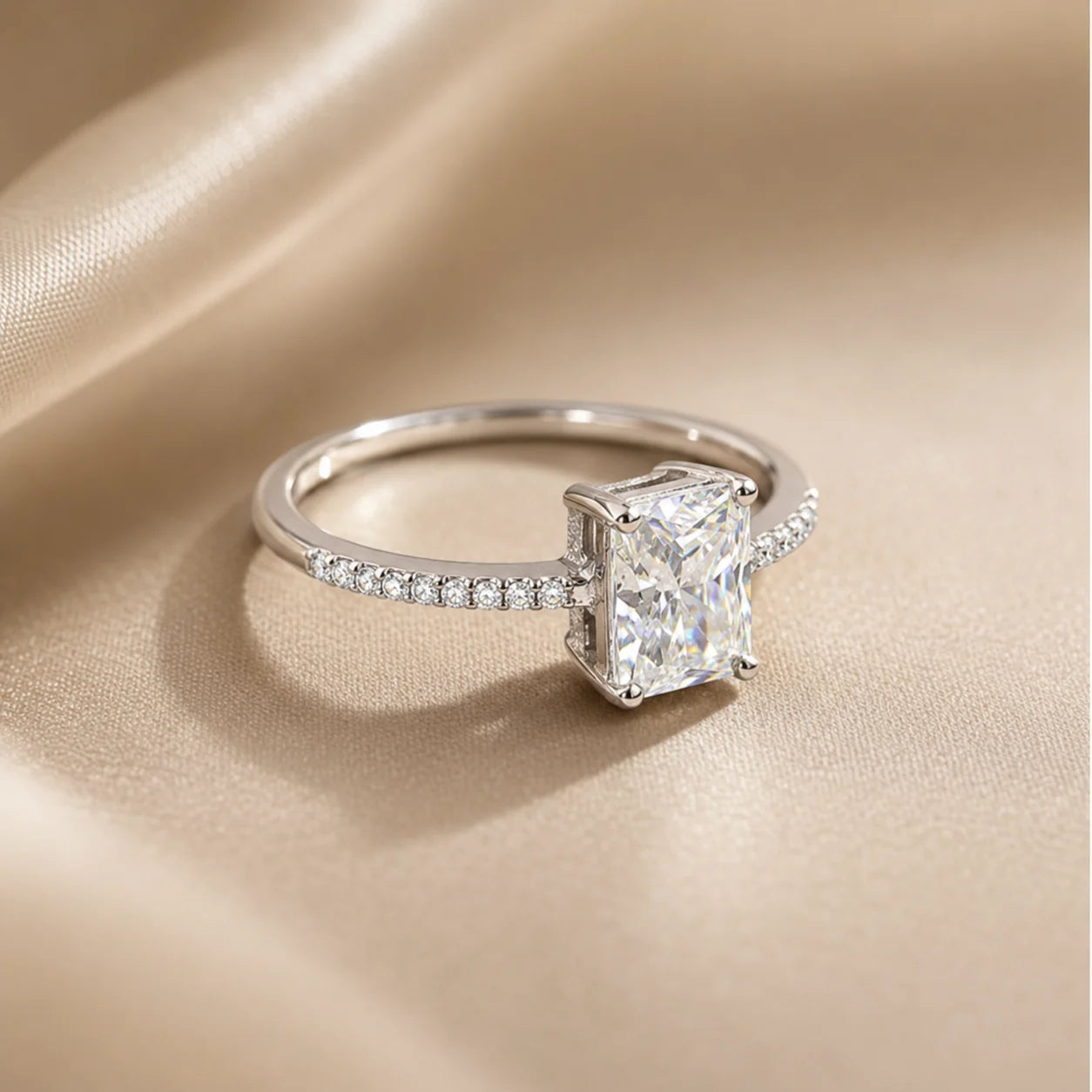Silver radiant-cut moissanite engagement ring displayed on soft champagne satin with a sparkling pavé band.