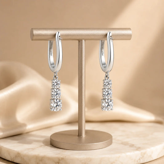 Three-stone sterling silver huggie drop earrings displayed on an elegant beige jewelry stand.