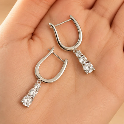 Three-stone sterling silver huggie drop earrings held in hand, showing the open hinged clasps and dangling design.