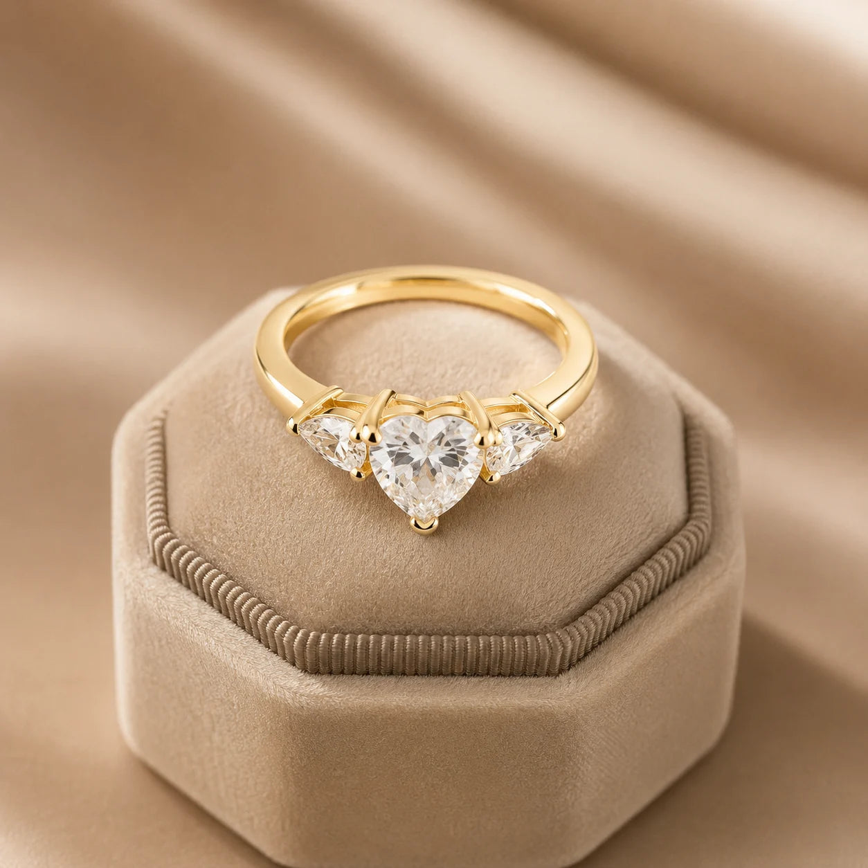 Yellow gold heart-cut three-stone moissanite engagement ring displayed on a beige velvet jewelry box with warm luxury lighting.