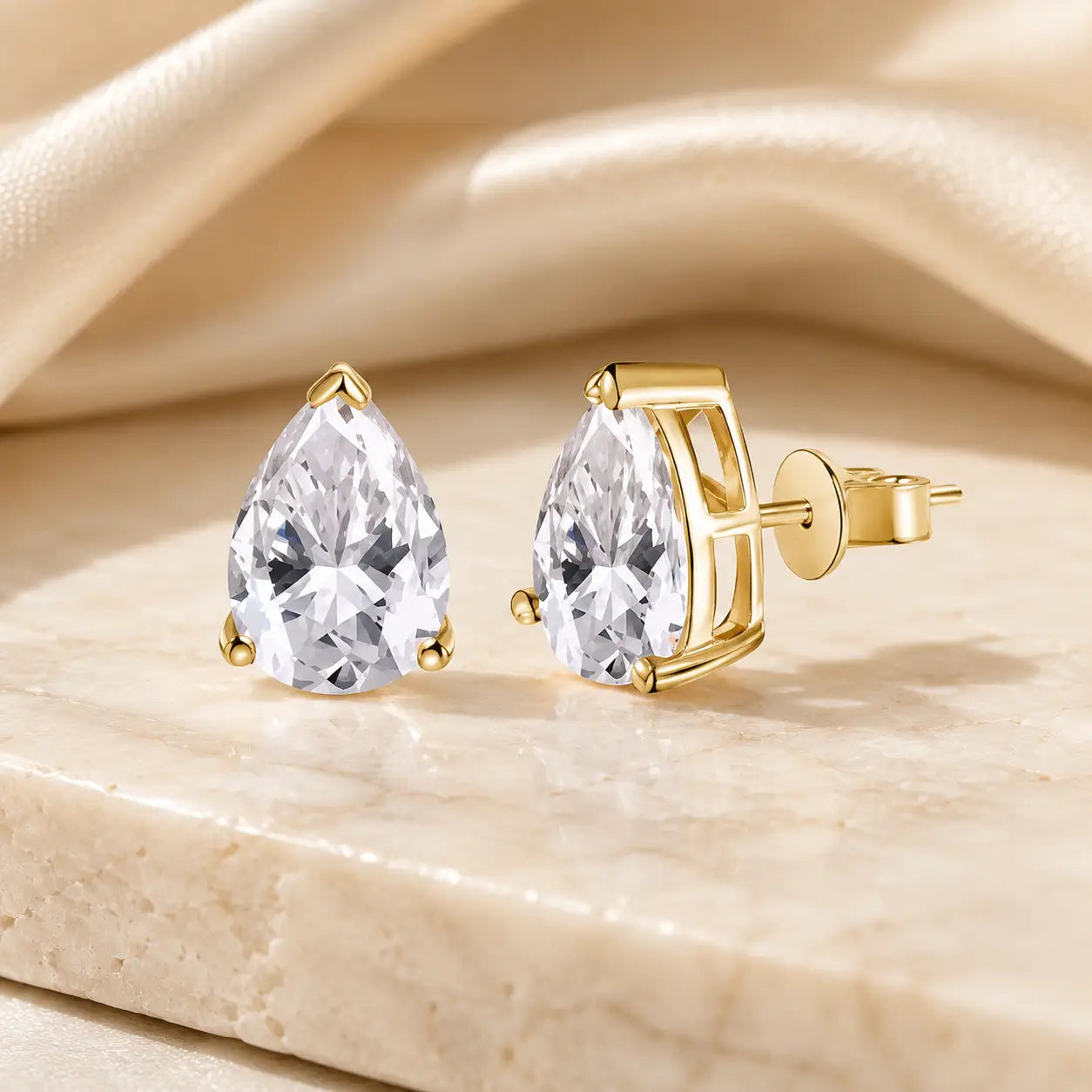 Yellow gold pear-cut moissanite stud earrings featuring brilliant pear-shaped stones on a soft beige display.